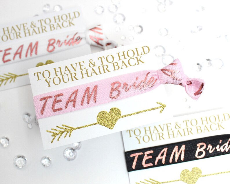 May include: Pink and black hair ties with gold glitter accents. The hair ties have the text "Team Bride" and "To Have & To Hold Your Hair Back" printed on them.