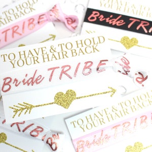 May include: White elastic hair ties with gold glitter arrows and hearts. The hair ties have the text "To Have & To Hold Your Hair Back" and "Bride Tribe" printed on them in pink.