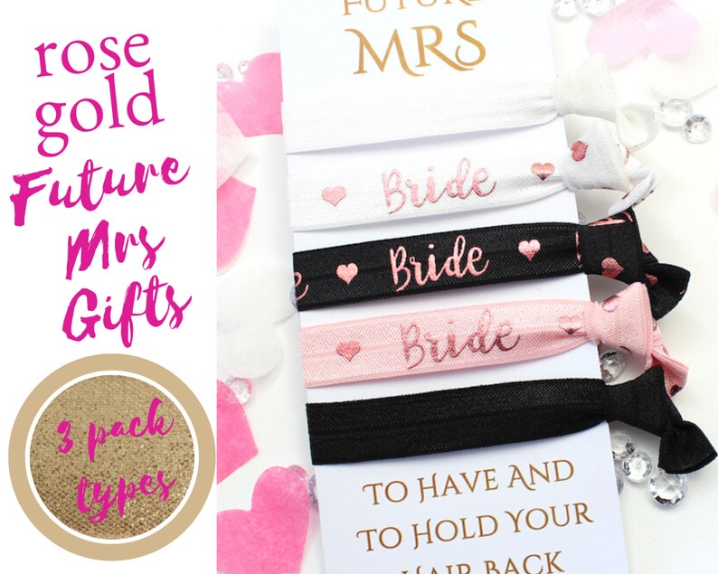 May include: Three pink and black elastic hair ties with the word "Bride" in rose gold lettering. The hair ties are packaged with a white card that reads "To have and to hold your hair back".