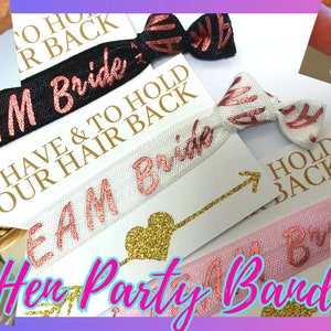 May include: A set of colorful elastic hair ties with text that says "Team Bride", "To Have & To Hold Your Hair Back", and "Hold Back". The hair ties are on a white background with gold glitter accents.