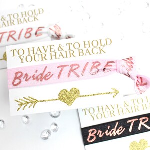 May include: Pink and black elastic hair ties with gold glitter lettering that reads "Bride Tribe" and "To Have & To Hold Your Hair Back".