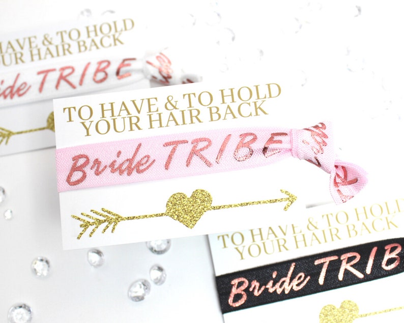 May include: Pink and black elastic hair ties with gold glitter lettering that reads "Bride Tribe" and "To Have & To Hold Your Hair Back".