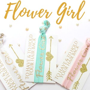 May include: Three white cards with gold glitter hearts and arrows, each with a different colored elastic hair tie attached. The cards read "To have & to hold your hair back" and "Flower Girl".