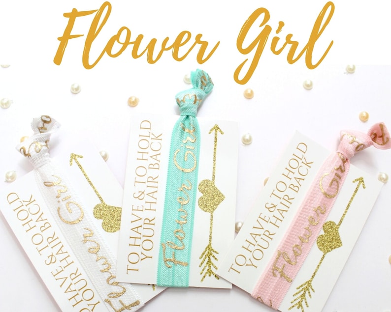 May include: Three white cards with gold glitter hearts and arrows, each with a different colored elastic hair tie attached. The cards read "To have & to hold your hair back" and "Flower Girl".