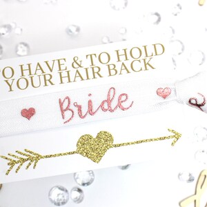 May include: White elastic hair tie with the words "To Have & To Hold Your Hair Back" and "Bride" in pink lettering. The hair tie is decorated with small pink hearts and a gold glitter arrow with a heart at the end.