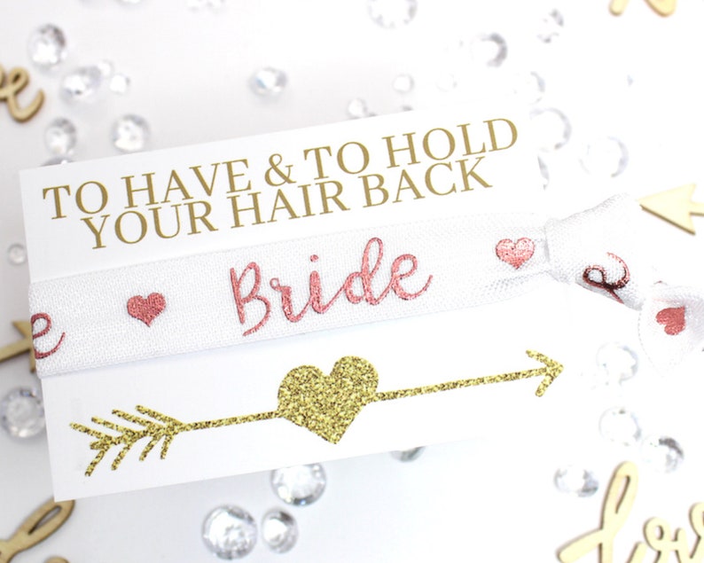 May include: White elastic hair tie with the words "To Have & To Hold Your Hair Back" and "Bride" in pink lettering. The hair tie is decorated with small pink hearts and a gold glitter arrow with a heart at the end.