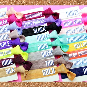 K&ouml;nnte beinhalten: Eine Sammlung bunter Haargummis mit Schleifenakzenten. Jedes Haarband hat eine andere Farbe, darunter Pfirsich, Burgunderrot und Schwarz. Die Namen der Farben sind auf wei&szlig;en Karten aufgedruckt. Der Hintergrund ist rosa.