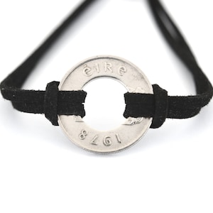 May include: A black suede cord bracelet featuring a silver coin with the word "Éire" and the year 1978. The coin is the focal point, with the cord threaded through its center.
