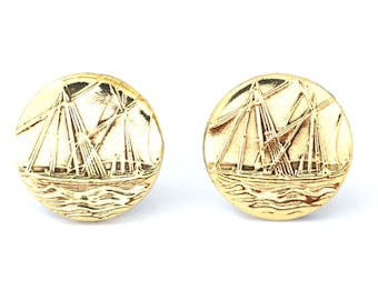 Könnte beinhalten: Zwei goldfarbene, runde Knöpfe mit einem detaillierten geprägten Design eines Segelboots auf dem Meer. Die Knöpfe haben eine glänzende Oberfläche und sind vor einem weißen Hintergrund platziert. Das Design zeigt die Masten, Segel und Wellen des Schiffs.