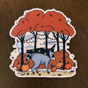 Könnte beinhalten: Ein gestanzter Aufkleber mit einem grauen Waschbären, der einen schwarzen Hexenhut mit gelben Sternen trägt und durch einen herbstlichen Wald mit orangefarbenen Bäumen, Kürbissen, kleinen weißen Geistern und Lichterketten läuft.