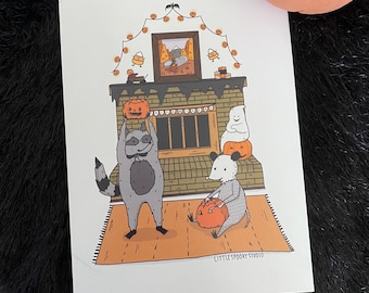 Puede incluir: Una ilustración de temática de Halloween con un mapache, un ratón y un fantasma sosteniendo calabazas talladas frente a una chimenea decorada con luces y maíz dulce. El texto "LITTLE SPOOKY STUDIO" está en la parte inferior de la imagen.