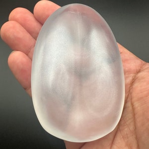 May include: A frosted, egg-shaped object with a smooth surface. The translucent material allows light to pass through, creating a soft, diffused appearance. The object is held in a hand against a dark background.