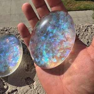 May include: Two oval-shaped, clear resin stones filled with iridescent, multi-colored foil flakes that reflect light in shades of blue, pink, and green. The smooth, polished surfaces of these decorative objects create a shimmering effect.