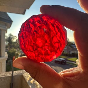 May include: A faceted, red crystal sphere is held up to the sunlight. The gem-like object has many flat surfaces that catch the light, creating a sparkling effect. The background shows a sunny outdoor scene with a house and a car.