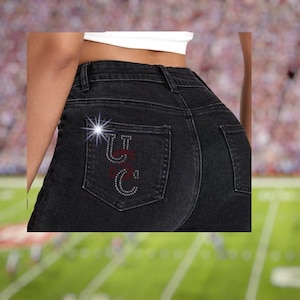 May include: Black denim jeans with a decorative design on the back pocket. The design features the letters "UC" in a combination of rhinestones and red lettering. The jeans are a dark wash and have a classic five-pocket design.