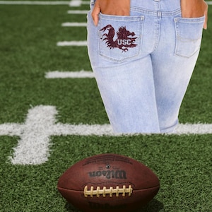 May include: Light blue denim jeans with a maroon USC logo on the back pocket. A brown leather football with white laces rests on a green field with white yard lines. The football is a Wilson brand.