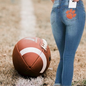 May include: A brown football with white stripes rests on the grass near a pair of blue jeans. The jeans have a sparkly orange paw print on the back pocket. The football is on a field with a white line.