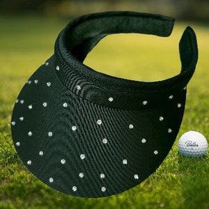 May include: A black golf visor with sparkling rhinestone accents. The visor sits on green grass next to a white golf ball. The visor has a curved brim and a comfortable headband. The golf ball has the word "Calloway" printed on it.