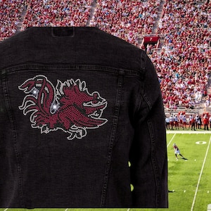 May include: Black denim jacket with a large, sparkling maroon and white design on the back. The design appears to be a stylized bird or creature. The jacket is set against a backdrop of a football stadium.