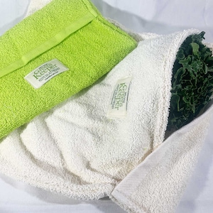 May include: Two white cotton produce savers with green trim and the text "Greens Keeper, Keeps Salads Fresh" on the front. One saver is folded and the other is holding a bunch of green leafy vegetables.