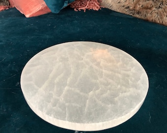 May include: A white, round selenite charging plate with a textured surface. The plate is on a teal blue background.