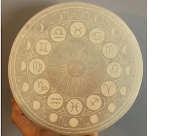 May include: A white circular stone with a zodiac wheel design. The wheel features the twelve zodiac signs and symbols, including Aries, Taurus, Gemini, Cancer, Leo, Virgo, Libra, Scorpio, Sagittarius, Capricorn, Aquarius, and Pisces. The center of the wheel features a sun with a face.