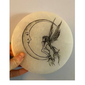 May include: A white circular stone with a black and white drawing of a fairy sitting on a crescent moon.