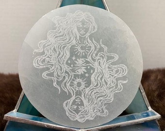 May include: A white selenite disc with an etched design of a woman with flowing hair and celestial symbols. The disc is on a blue stained glass stand.