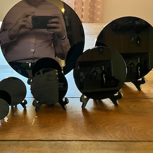 Peut inclure: Cinq miroirs circulaires noirs de tailles diff&eacute;rentes sont pr&eacute;sent&eacute;s sur de petits supports noirs. Les miroirs sont dispos&eacute;s en rang&eacute;e sur une surface en bois.