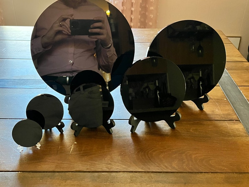 Peut inclure: Cinq miroirs circulaires noirs de tailles diff&eacute;rentes sont pr&eacute;sent&eacute;s sur de petits supports noirs. Les miroirs sont dispos&eacute;s en rang&eacute;e sur une surface en bois.