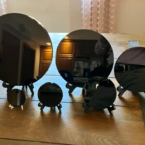 Peut inclure: Cinq miroirs circulaires noirs de tailles diff&eacute;rentes sont pr&eacute;sent&eacute;s sur des supports en bois. Les miroirs sont dispos&eacute;s en demi-cercle sur une table en bois.
