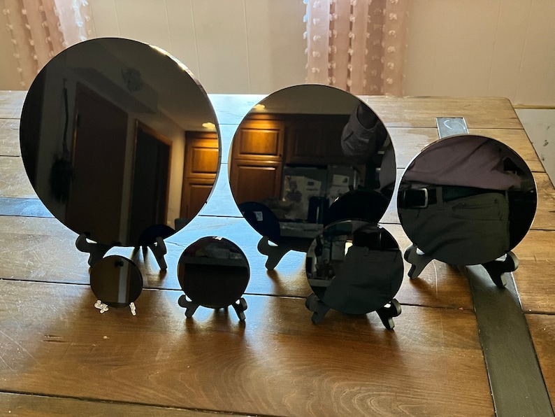 Peut inclure: Cinq miroirs circulaires noirs de tailles diff&eacute;rentes sont pr&eacute;sent&eacute;s sur des supports en bois. Les miroirs sont dispos&eacute;s en demi-cercle sur une table en bois.