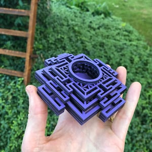 Puede incluir: Un objeto cuadrado de color morado, de varias capas, con un agujero circular en el centro. El objeto está hecho de madera y tiene un patrón geométrico.