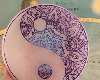 May include: A white, round Yin Yang stone with intricate mandala designs in shades of blue and purple. The design is hand-drawn, with a detailed floral pattern. The background features a vintage map.