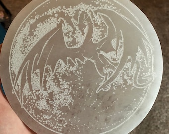 May include: A white ceramic coaster with a raised design of a dragon flying in front of a moon. The moon is filled with small white dots.