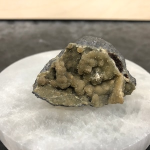 May include: A dark gray mineral specimen with a cluster of light green, textured formations. The mineral sits on a white, circular, polished surface, with a dark background.