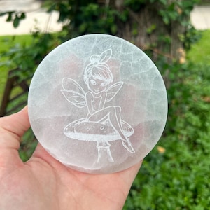May include: A round, white selenite crystal plate with a detailed etched image of a fairy sitting on a mushroom. The fairy has wings and is wearing a dress. The plate is held in a hand.