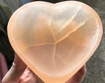 May include: A heart-shaped bowl made of light pink selenite crystal. The bowl has a smooth, polished surface and a natural crack running through the center.