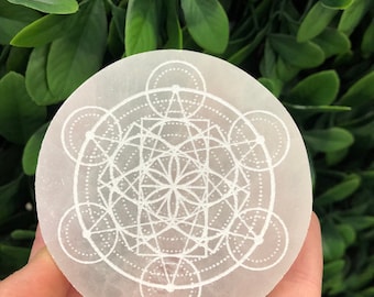 May include: White selenite crystal disc with a geometric design etched into the surface. The design features a flower of life pattern with circles and lines.