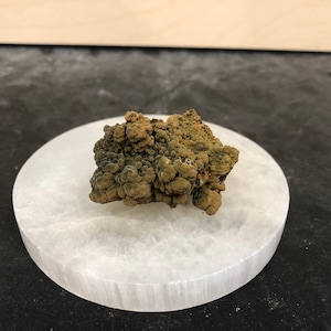 May include: A small, irregularly shaped, brown and green rock on a white, round, textured surface.