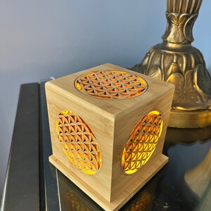 May include: A wooden cube with a geometric pattern carved into each side. The cube is illuminated from within, creating a warm glow. The pattern is known as the Flower of Life.