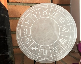 May include: A white circular zodiac wheel with the astrological signs and symbols for each of the twelve zodiac signs. The wheel is on a black metal stand.