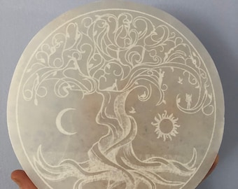 May include: White selenite disc with an etched tree of life design. The tree has intricate branches and roots, and there is a crescent moon and sun on either side of the trunk.