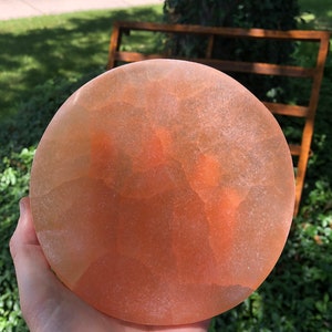 May include: A round, light orange, translucent, natural selenite crystal plate. The plate has a smooth, polished surface and a slightly rough, natural edge.