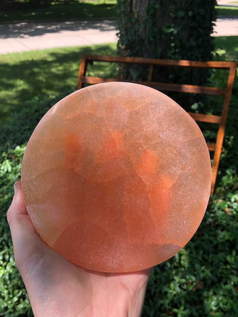 May include: A round, light orange, translucent, natural selenite crystal plate. The plate has a smooth, polished surface and a slightly rough, natural edge.