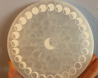 May include: A white, round, frosted glass plate with a moon phase design and a sun with a crescent moon in the center.