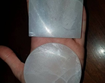 May include: Two selenite charging plates, one square and one round, in a person's hand. The plates are white and have a smooth, polished surface.