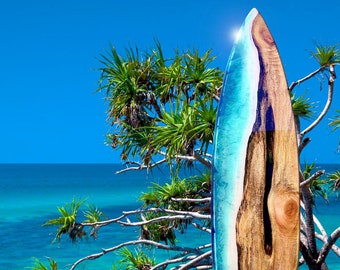 Op de afbeelding: Een surfboard met een blauw en wit harsontwerp op de bovenste helft en een natuurlijke houtnerf op de onderste helft. Het surfboard staat rechtop op een grasveld met een strand en de oceaan op de achtergrond.