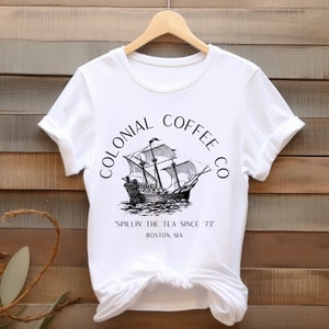 May include: White t-shirt featuring a black and white illustration of a tall ship with the text "COLONIAL COFFEE CO" arched above the ship. Below the ship is the text "'SPILLIN' THE TEA SINCE '73" and "BOSTON, MA".