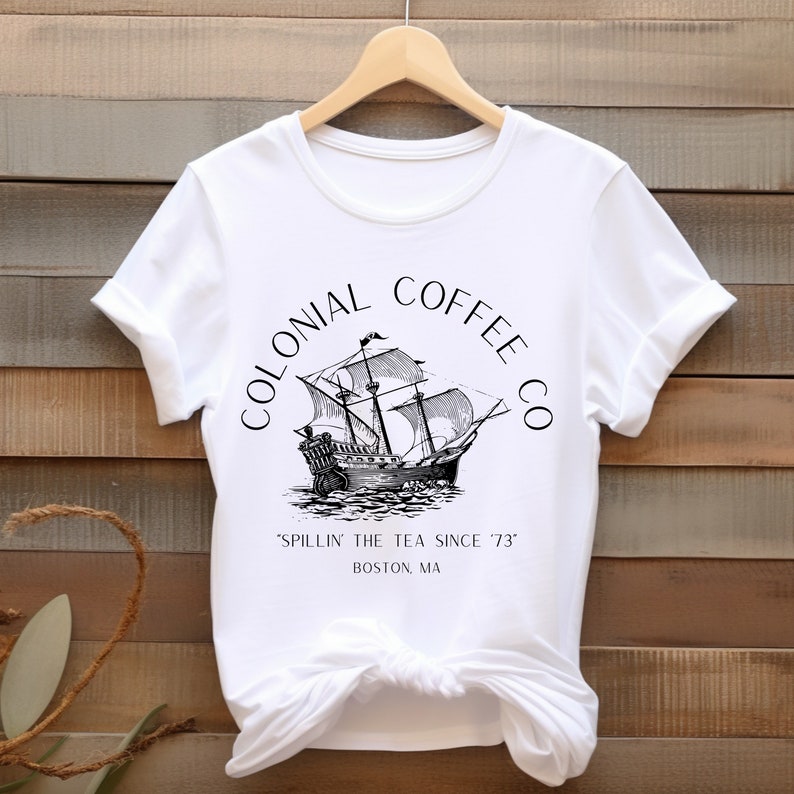 May include: White t-shirt featuring a black and white illustration of a tall ship with the text "COLONIAL COFFEE CO" arched above the ship. Below the ship is the text "'SPILLIN' THE TEA SINCE '73" and "BOSTON, MA".